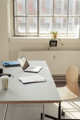 Laptop, documents and a paper coffee cup on a white desk in the interior of a modern bright office with large window, radiator and a wooden chair on wheels